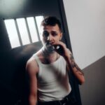 Adult man in tank top smokes cigarette near barred window, evoking prison theme.