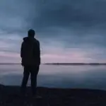 A solitary silhouette of a man in a jacket gazing at a lake during a peaceful sunset, creating a serene atmosphere.